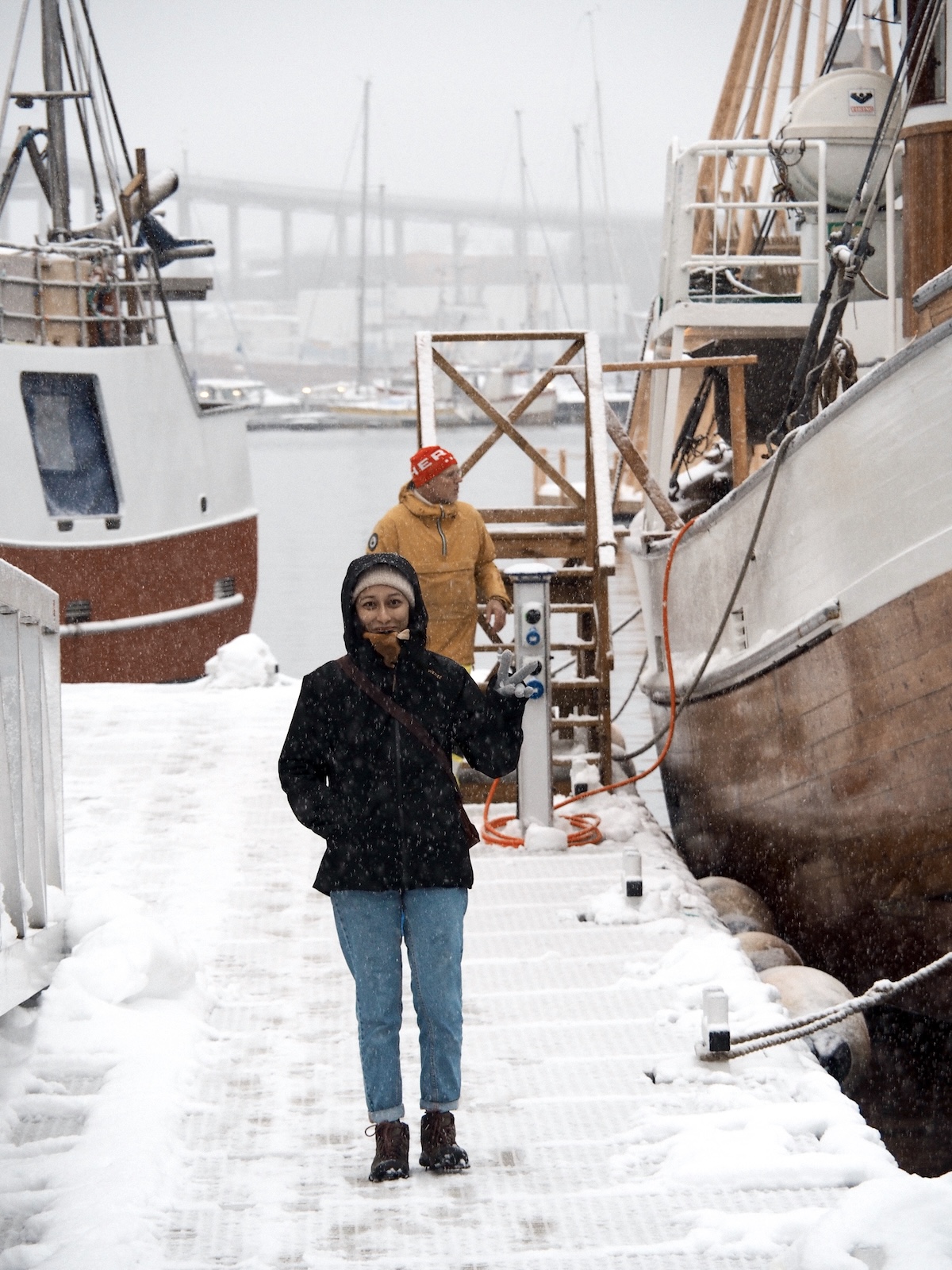 Person walking along a snowy marina dock in Tromsø, Norway, past fishing boats during snowfall