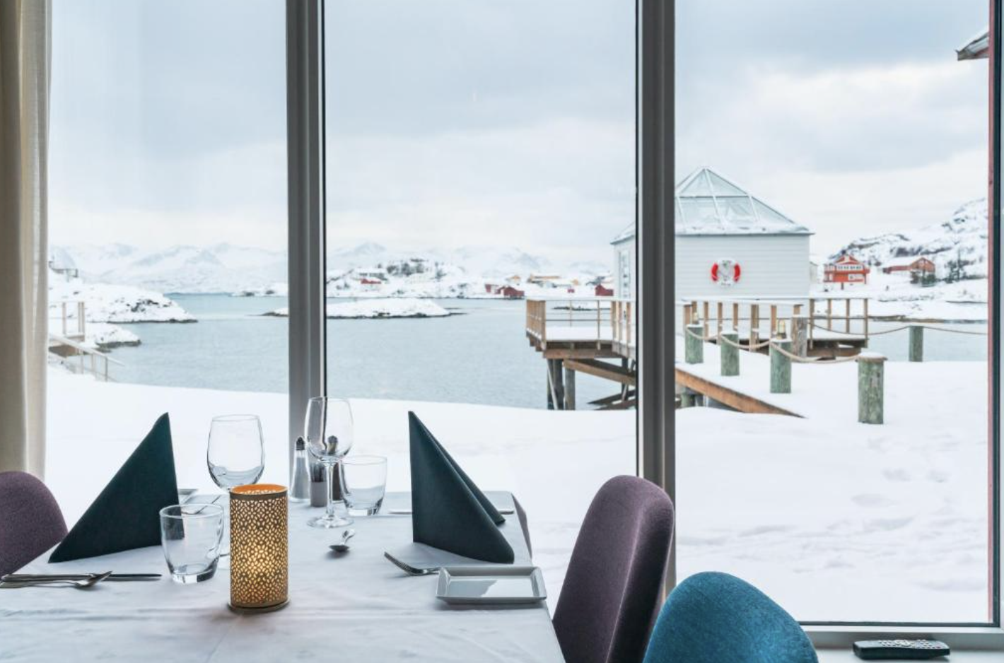 Set restaurant table with candlelight facing floor-to-ceiling windows overlooking a snowy Sommarøy waterfront in Norway